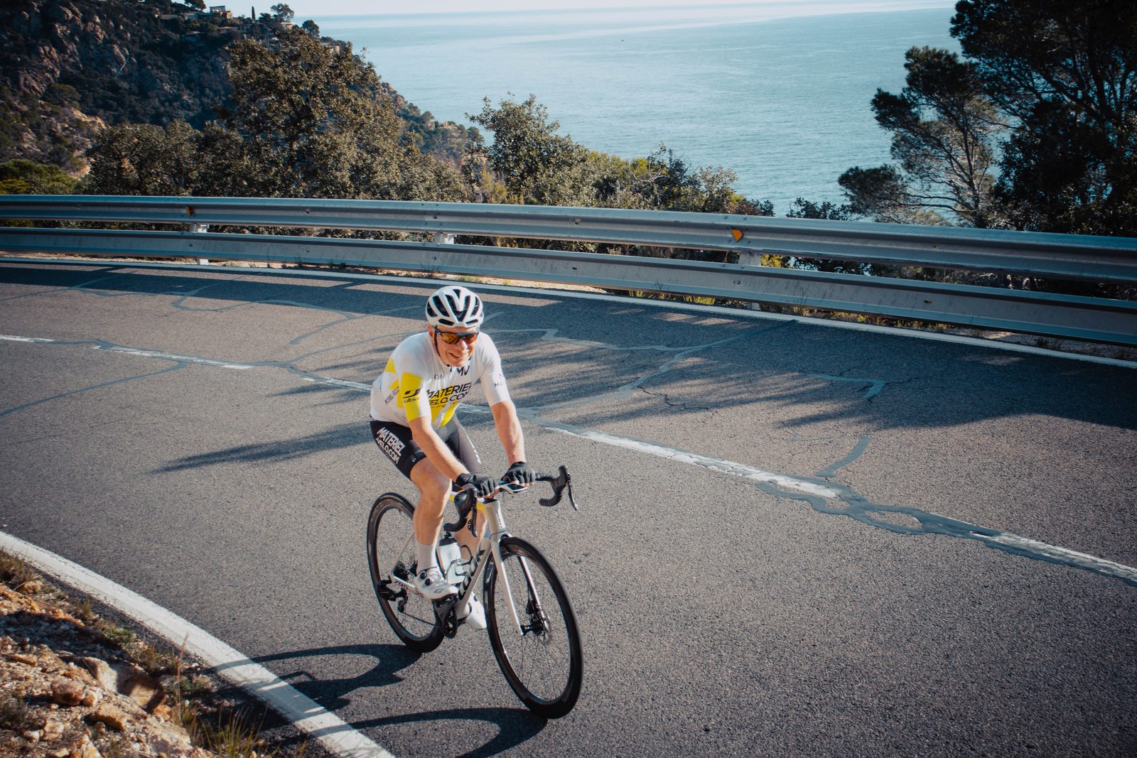 Un cycliste en maillot Materiel-velo.com sourit dans un virage d’une route côtière, la mer et les pins en contrebas, sur la Costa Brava, pas sur la Riviera italienne.