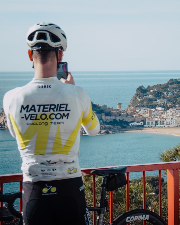 Un cycliste en maillot Materiel-velo.com, de dos, photographie avec son téléphone une baie de la Costa Brava, un village blanc et une tour sur le promontoire.