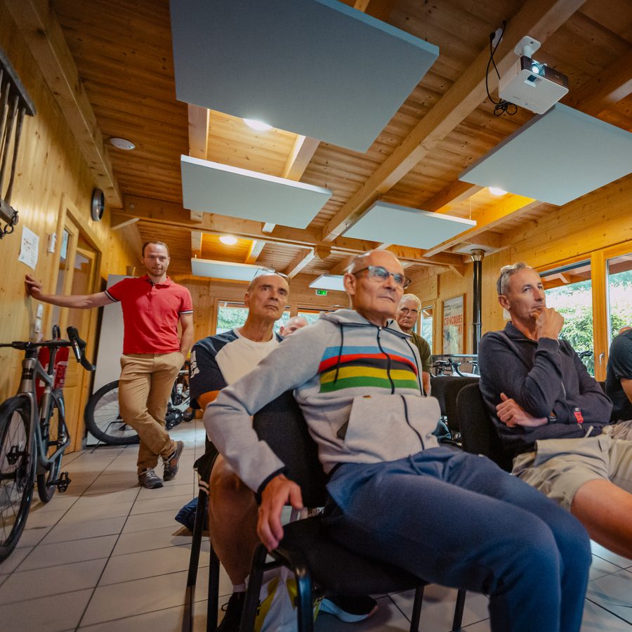 Un atelier coaching dans une salle boisée : un encadrant en polo rouge, debout contre le mur, face à des cyclistes assis qui l’écoutent.
