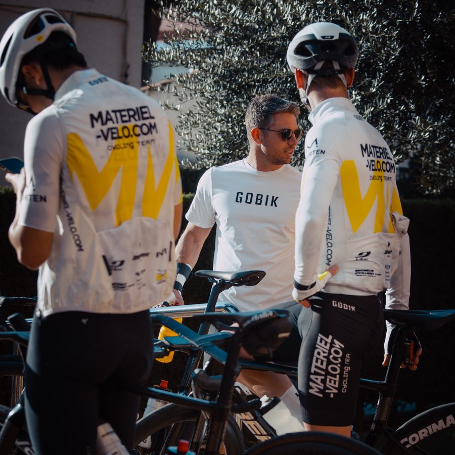 Un encadrant en tee-shirt blanc parle avec deux cyclistes en maillot Materiel-velo.com, vus de dos, au milieu des vélos.