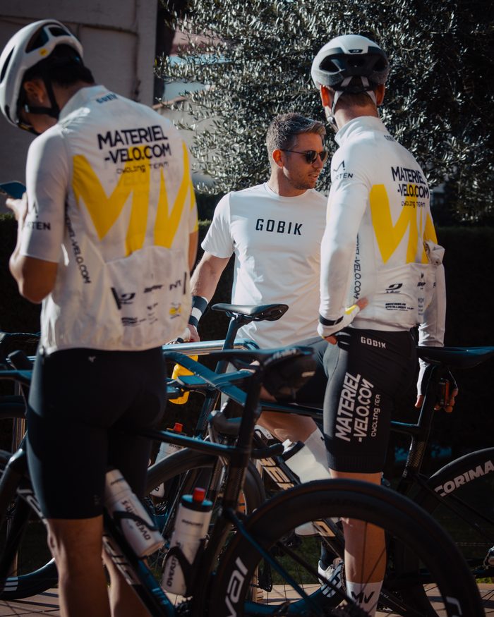 Un encadrant en tee-shirt blanc discute avec deux cyclistes en maillot Materiel-velo.com, vus de dos, au milieu des vélos.