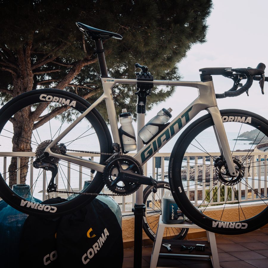 Un vélo de course sur un pied d’atelier, sur une terrasse au-dessus de la mer, des pins parasols derrière la balustrade.