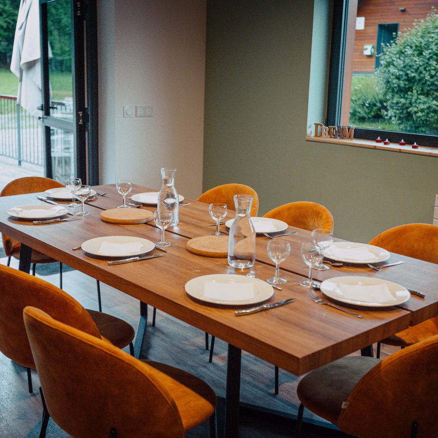 Une salle à manger, une longue table en bois dressée pour le repas, des chaises orange et une baie vitrée ouverte sur la verdure.