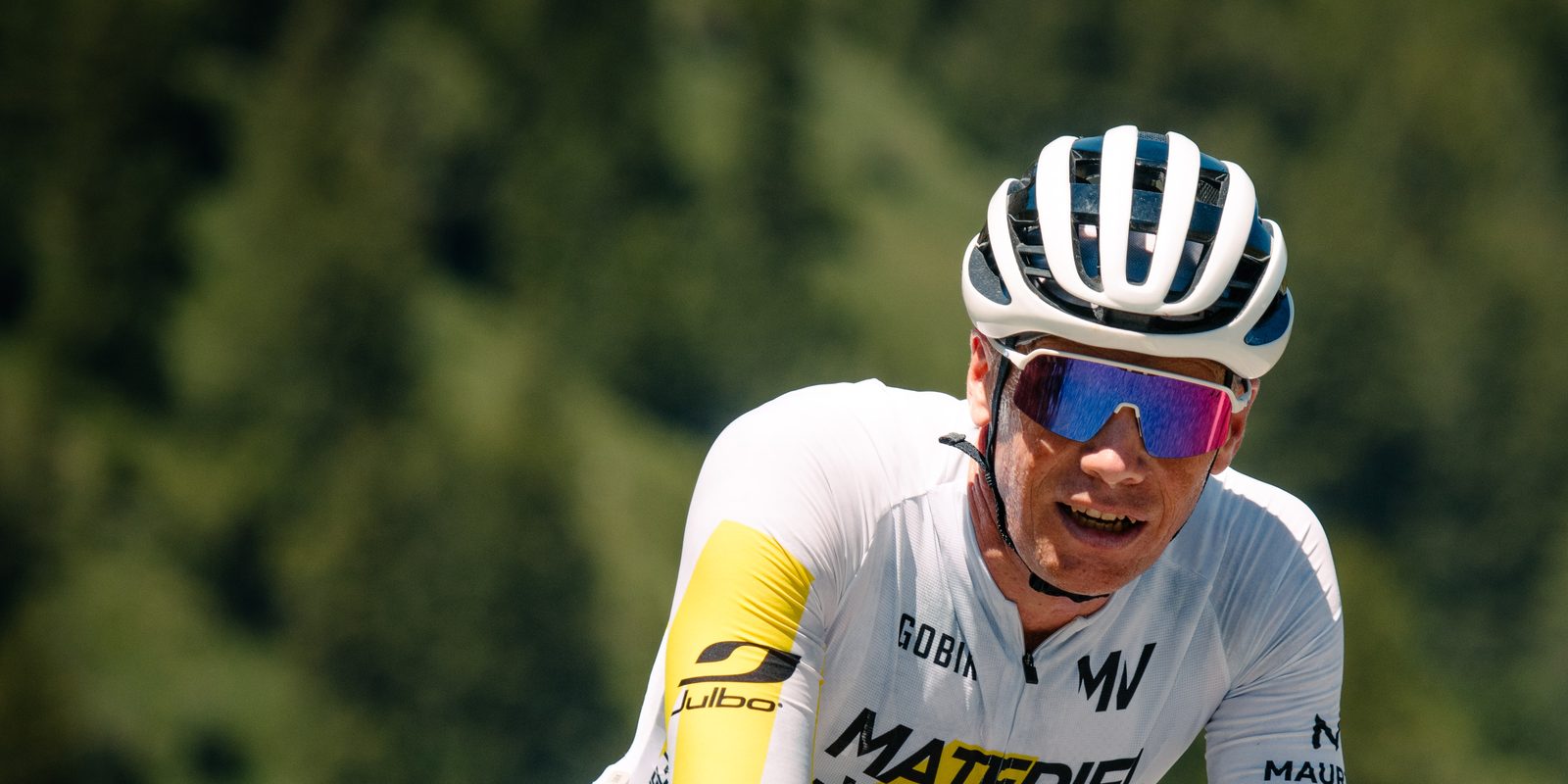 Un cycliste en maillot blanc et jaune Materiel-velo.com, casque blanc et lunettes, en plein effort sur une route de montagne, la forêt floue derrière lui.