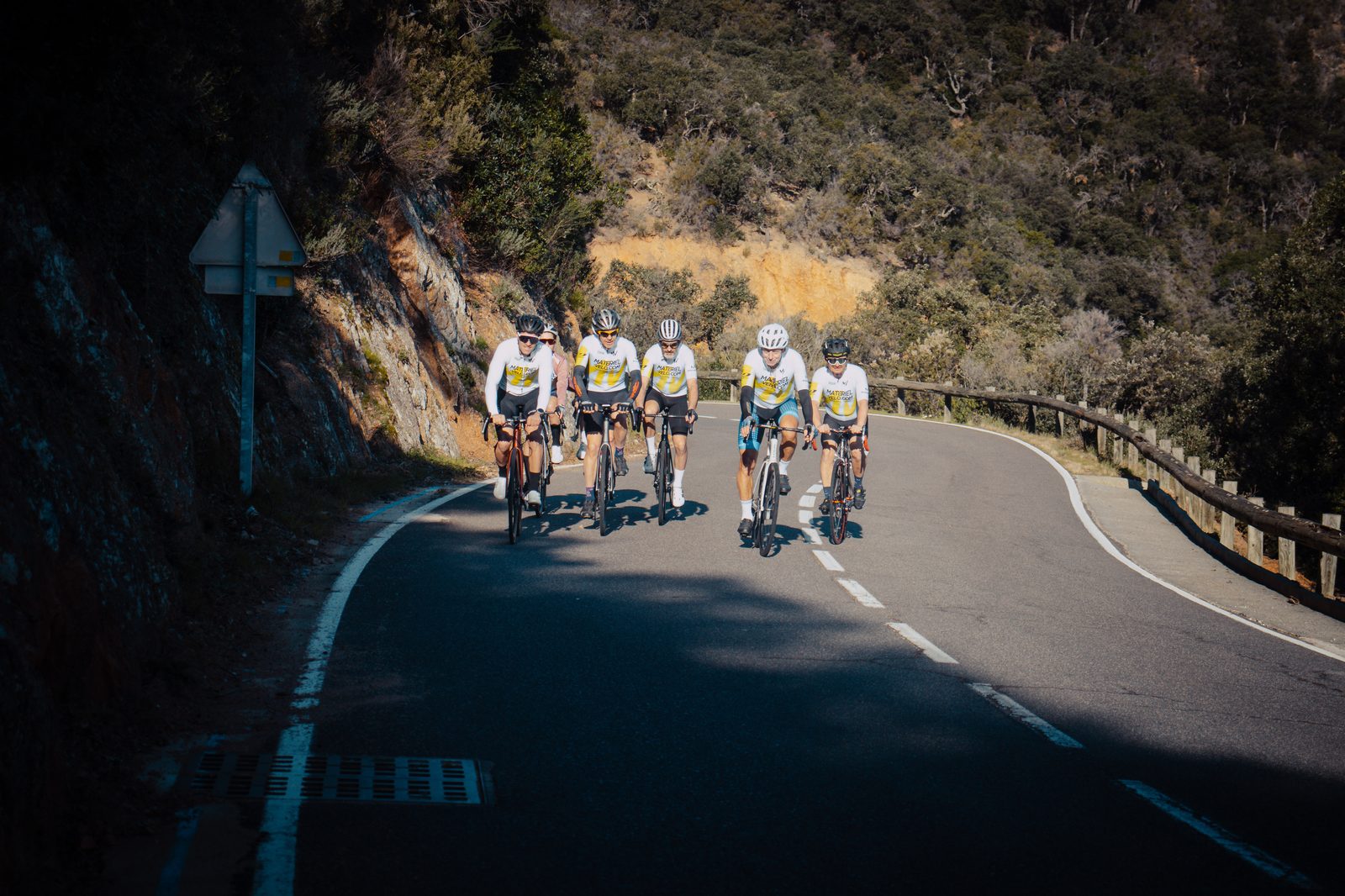 Six cyclistes arrivent de face sur une route de la Costa Brava, cinq en tenue blanche et jaune de l’équipe et un en haut rose, paroi rocheuse ocre à gauche et glissière à droite.