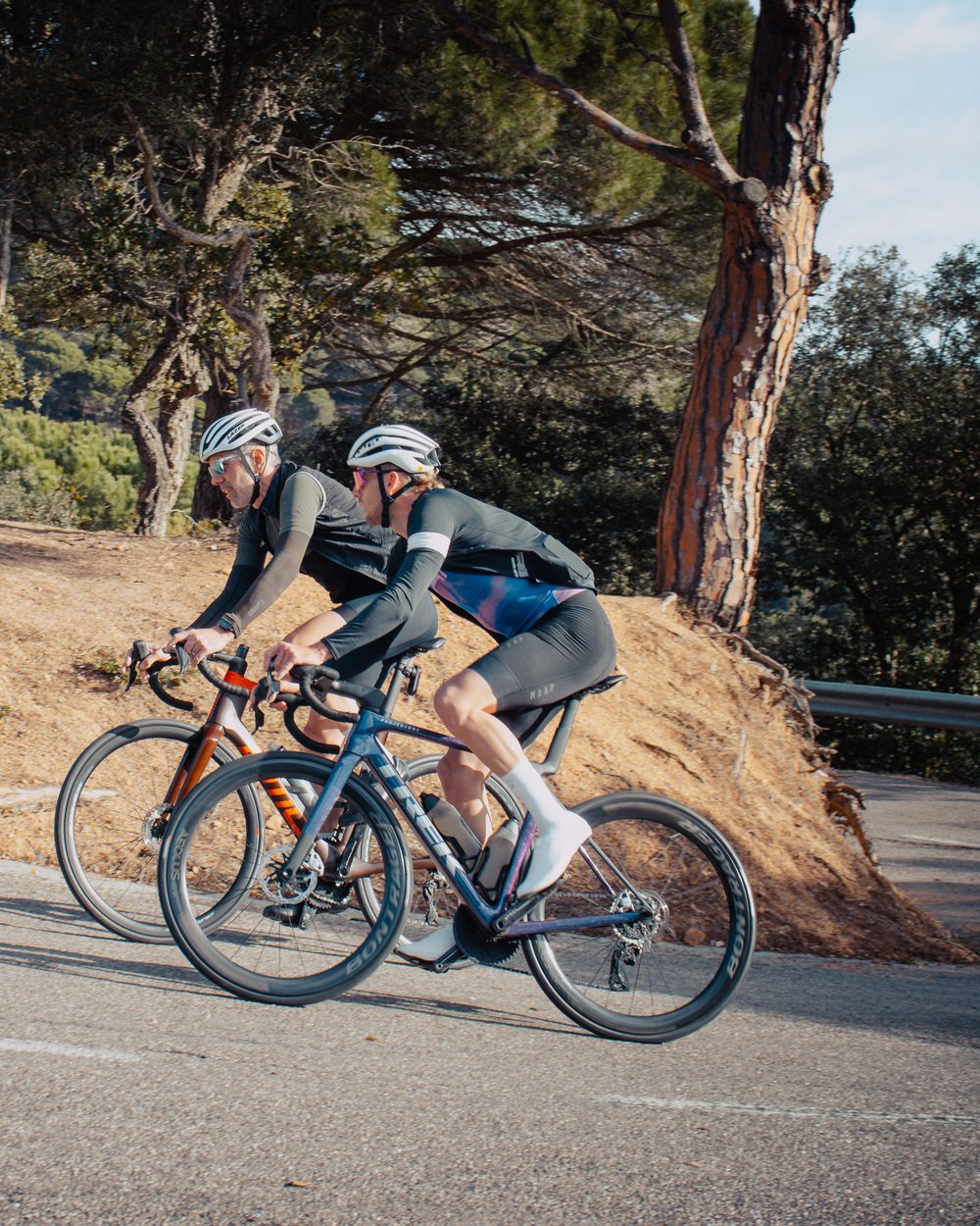 Deux cyclistes de profil sur une route de la Costa Brava, en tenue sombre, un talus ocre et de grands pins derrière eux.