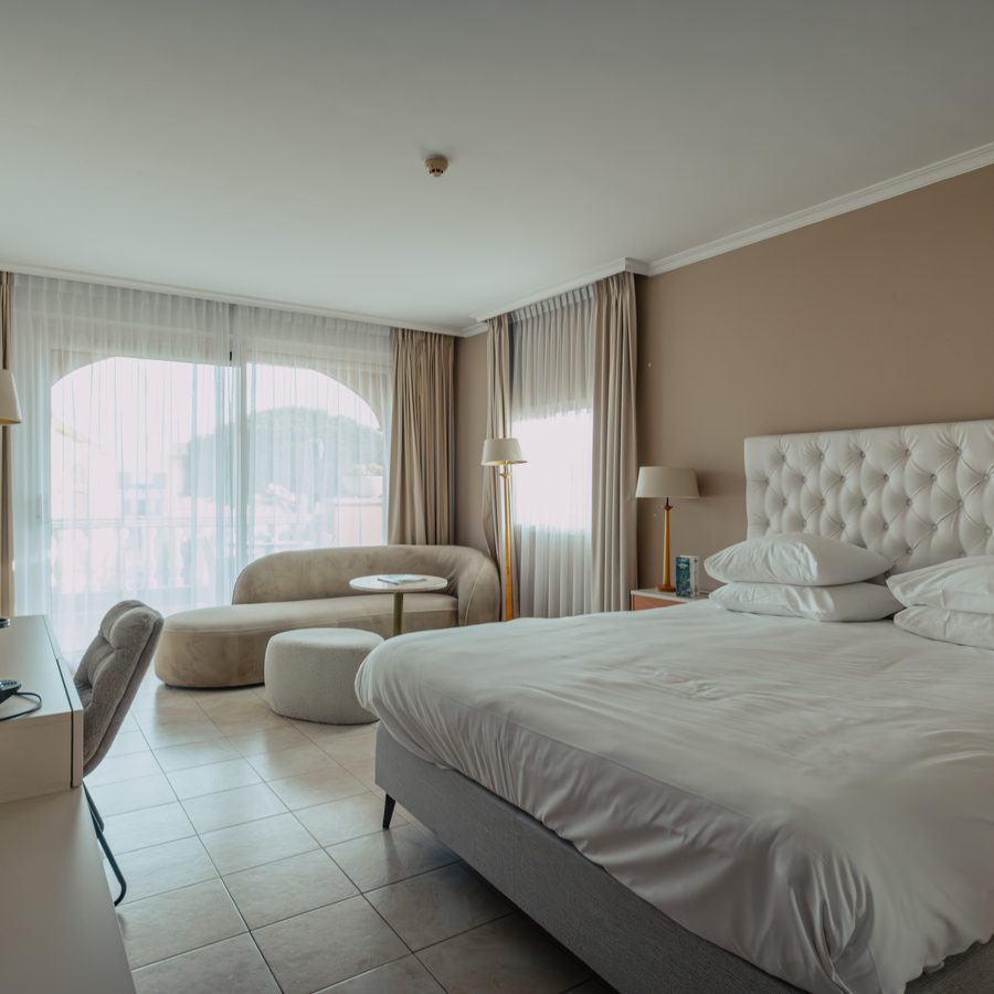 Une chambre de l’hôtel, une méridienne et un pouf devant la porte-fenêtre aux voilages blancs, le grand lit à droite.