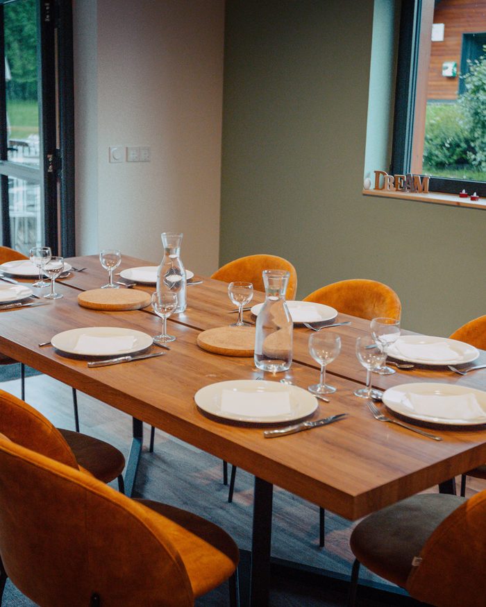 Une table en bois dressée pour le repas, assiettes, verres et carafes, entourée de chaises orange.