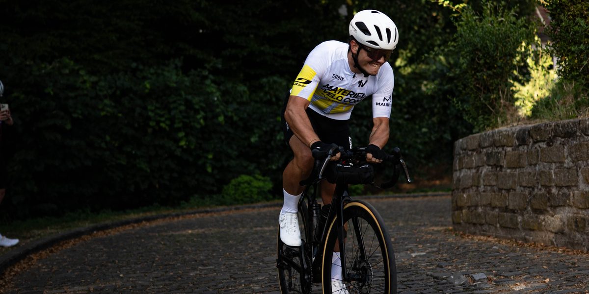 Un cycliste en maillot blanc et jaune Materiel-velo.com grimpe une route pavée en virage, le long d’un muret de pierre, sous de grands arbres.
