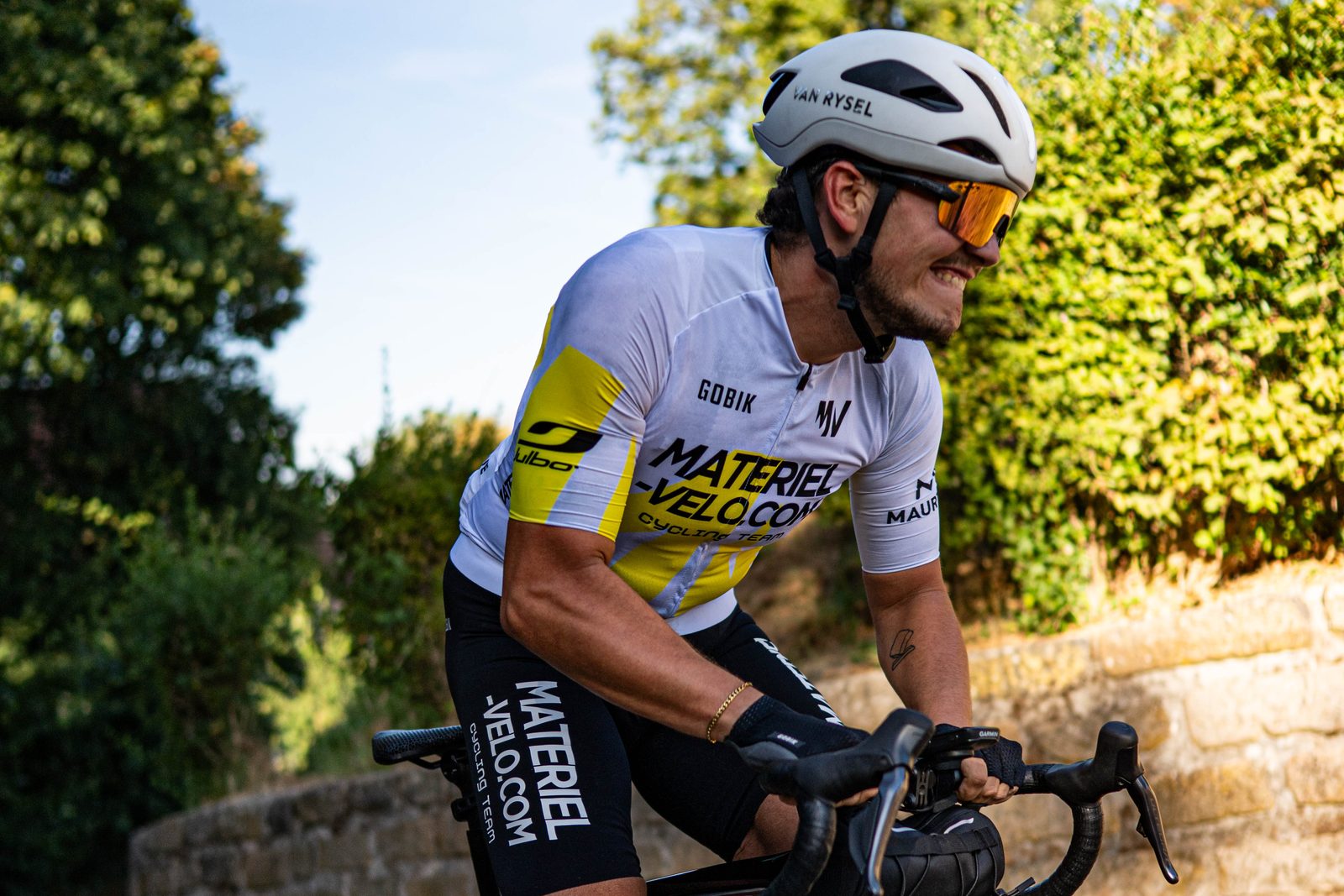 Un cycliste en maillot blanc et jaune Materiel-velo.com, les dents serrées, devant un muret de pierre et des feuillages.