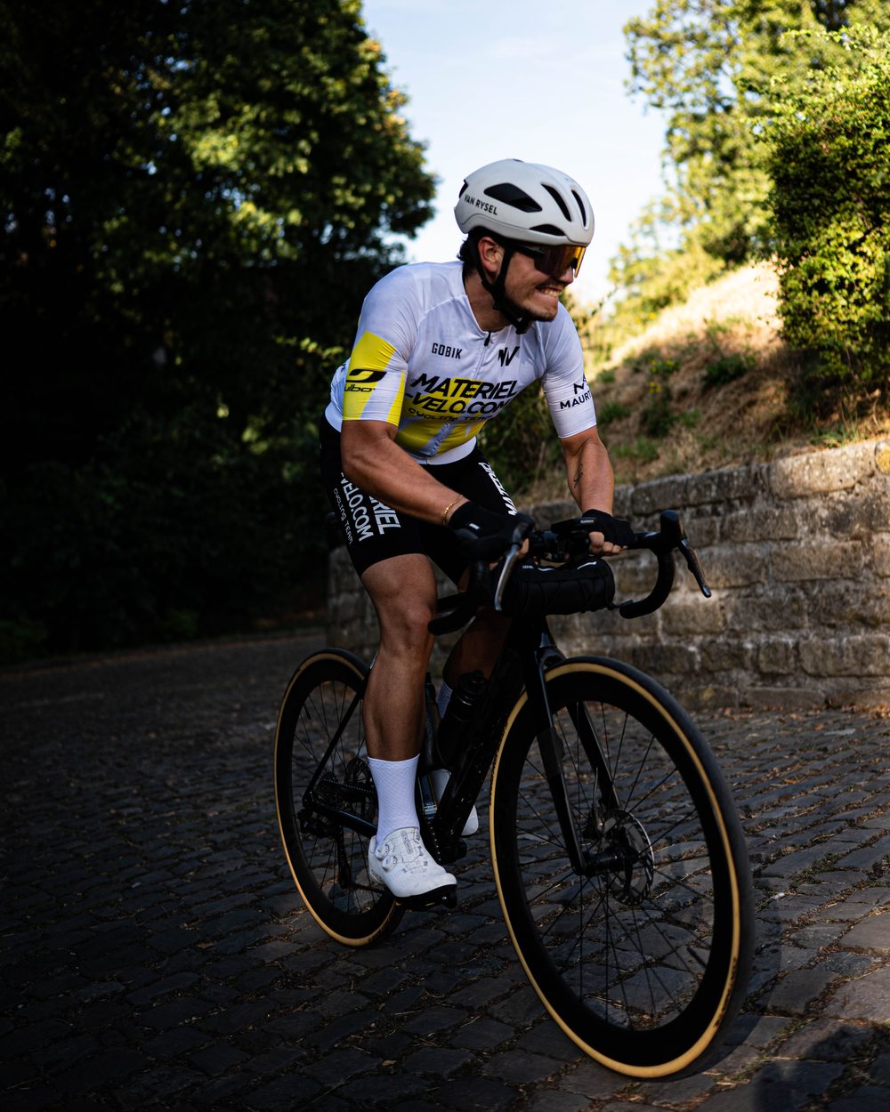 Un cycliste en maillot Materiel-velo.com en danseuse sur les pavés, un mur de pierre à sa droite.
