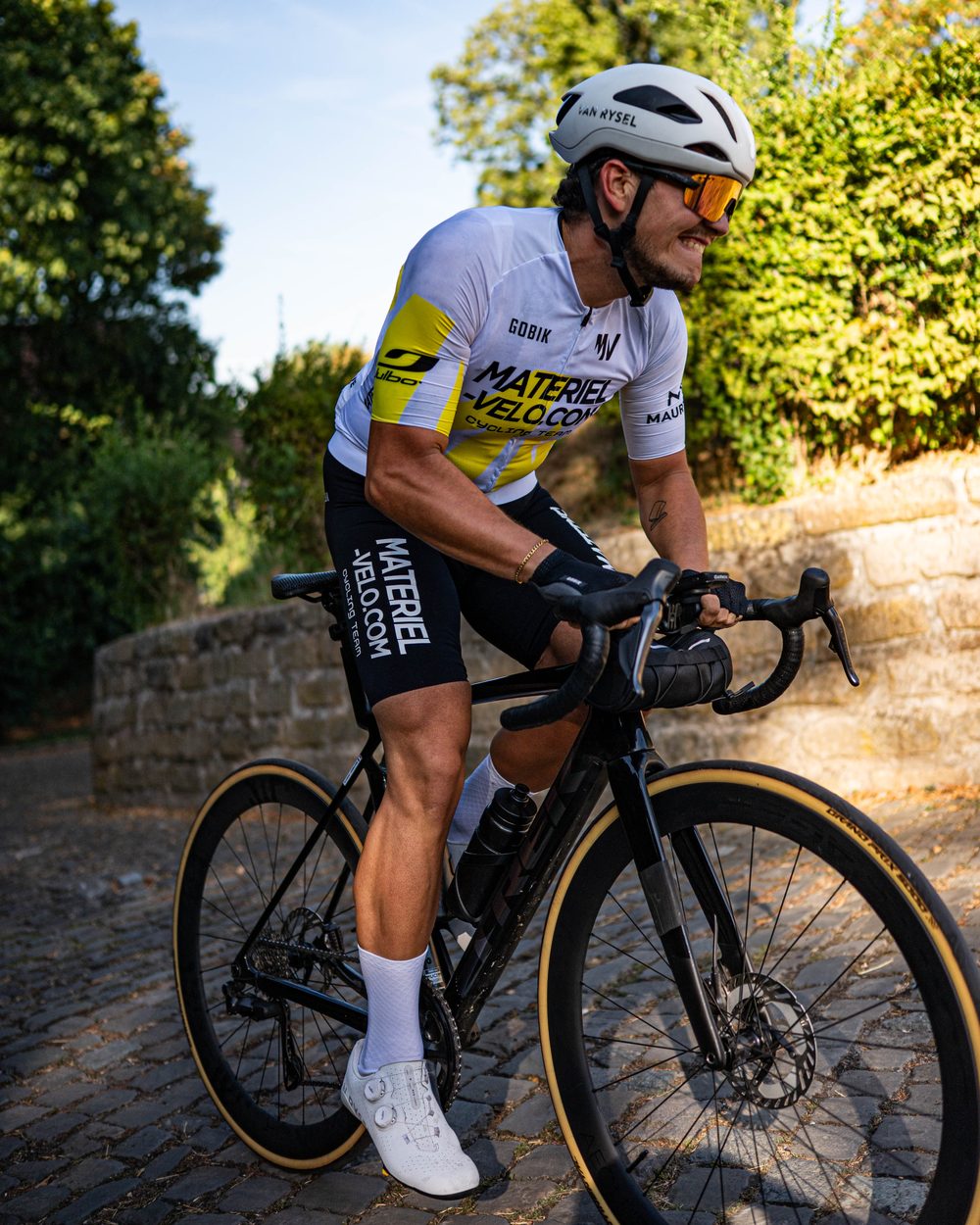 Un cycliste en maillot Materiel-velo.com de trois quarts sur une route pavée, en fin de journée.