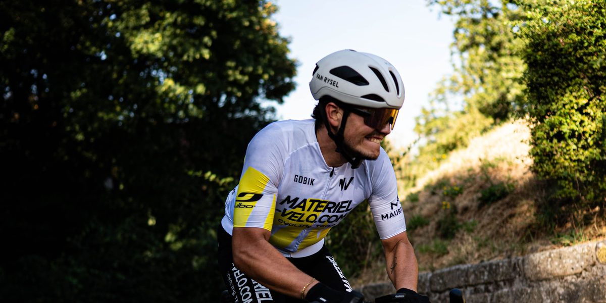 Un cycliste en maillot blanc et jaune Materiel-velo.com et casque blanc, souriant, sur une montée pavée bordée d’un muret de pierre.