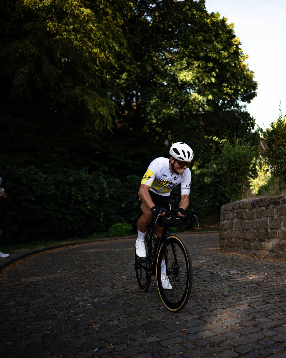 Un cycliste en maillot Materiel-velo.com monte une côte pavée bordée d’arbres et d’un mur de pierre.