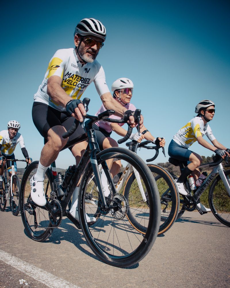Quatre cyclistes roulent vers l’objectif sur une route ensoleillée, celui de devant en pied dans la tenue de l’équipe, ciel bleu.